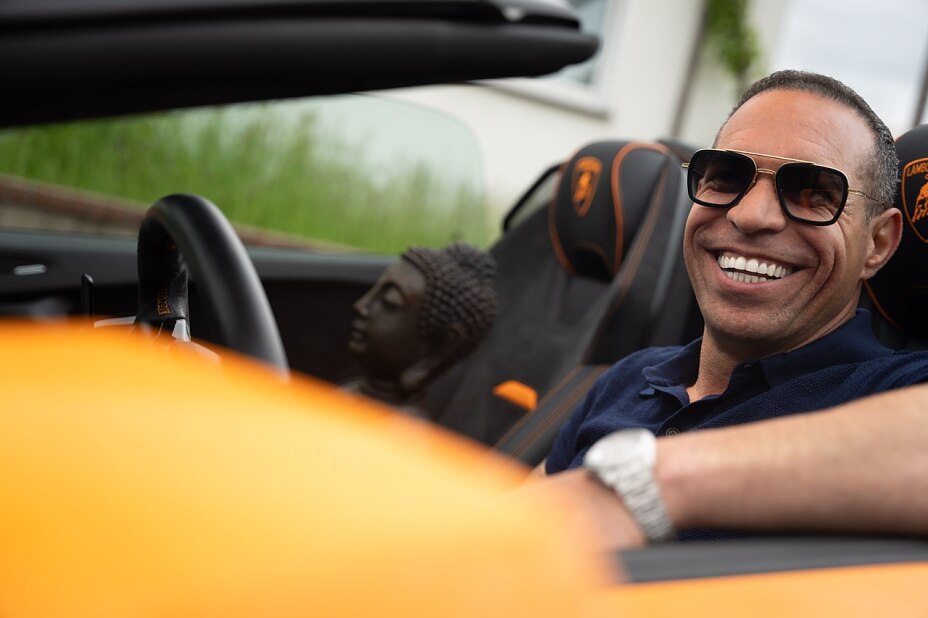 Eine Nahaufnahme von Marc Galal, der lachend und mit einer Sonnenbrille auf der Nase am Steuer eines orangefarbenen Cabrio-Sportwagens sitzt. Auf dem Beifahrersitz im Hintergrund steht eine dunkle Buddha-Kopf-Statue.