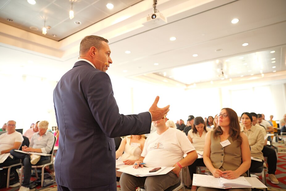 Der Verkaufstrainer Marc Galal steht im Profil in einem Seminarraum und spricht mit erhobener, gestikulierender Hand zu einer Gruppe aufmerksam zuhörender Teilnehmer, die Seminarunterlagen auf dem Schoß haben.