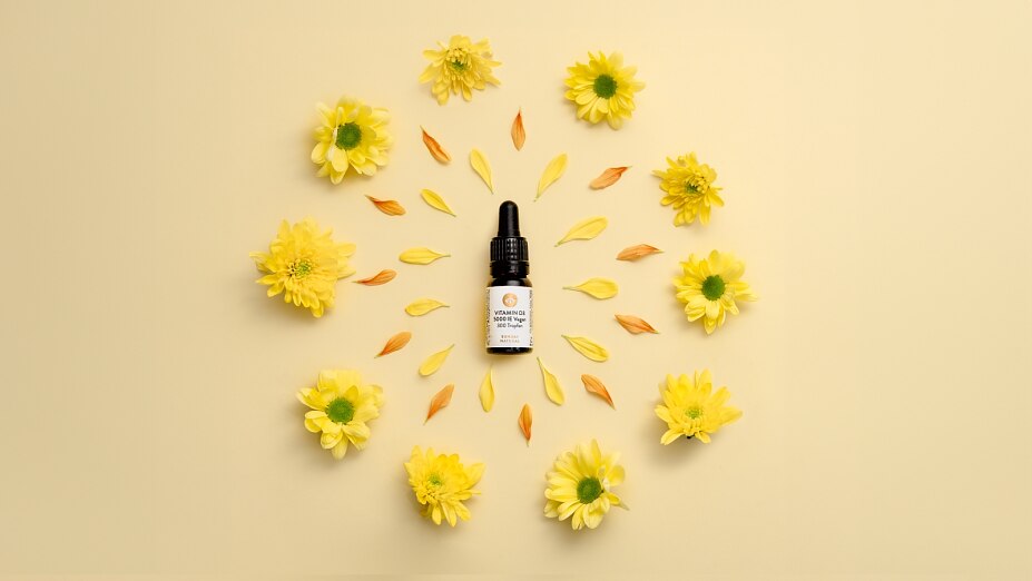 Eine Flasche Vitamin D3 5000 IE von Sunday Natural, die in der Mitte eines Kreises aus gelben Chrysanthemen-Blüten und Blütenblättern auf hellem Hintergrund liegt.