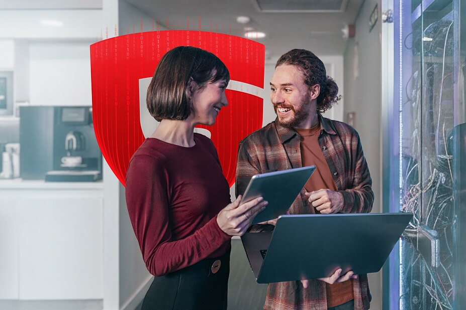 Ein Mann und eine Frau stehen lächelnd vor einem Serverschrank und arbeiten gemeinsam an einem Laptop; im Hintergrund ein rotes Schutzschild-Symbol.