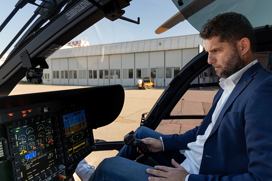 Pilot mit blauem Jacket sitzt in Hubschrauber Cockpit. Im Hintergrund eine Halle mit weissen Toren.