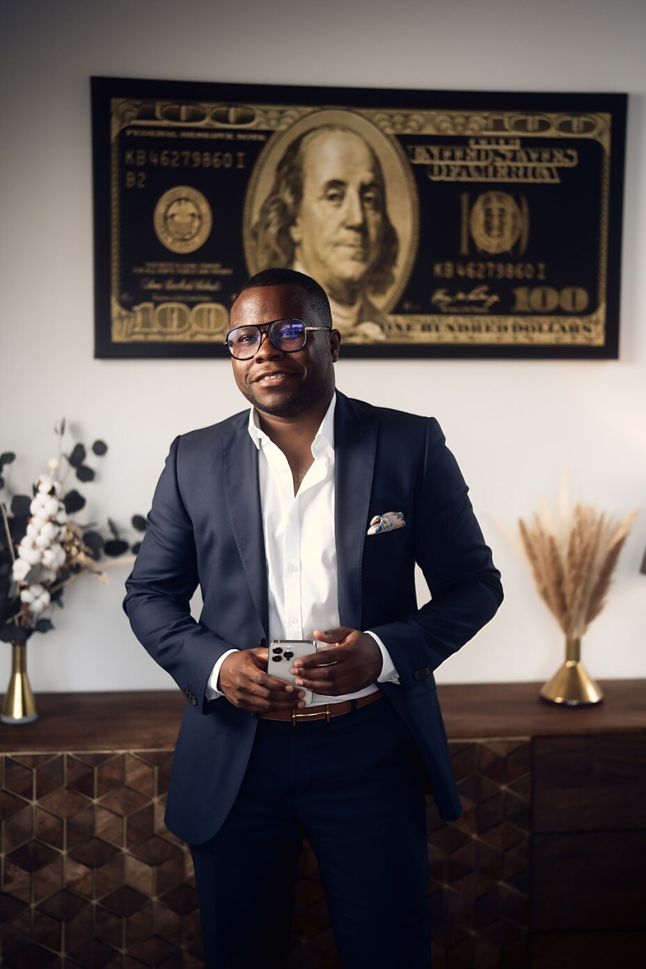 Portrait von Alvin Lisebushira in einem eleganten blauen Anzug vor einem modernen Wandbild, das eine 100-Dollar-Note zeigt