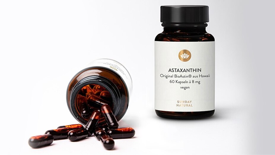 Eine stehende und eine liegende braune Glasflasche mit Astaxanthin-Kapseln von Sunday Natural. Mehrere dunkelrote Kapseln liegen verstreut vor der Öffnung der liegenden Flasche.