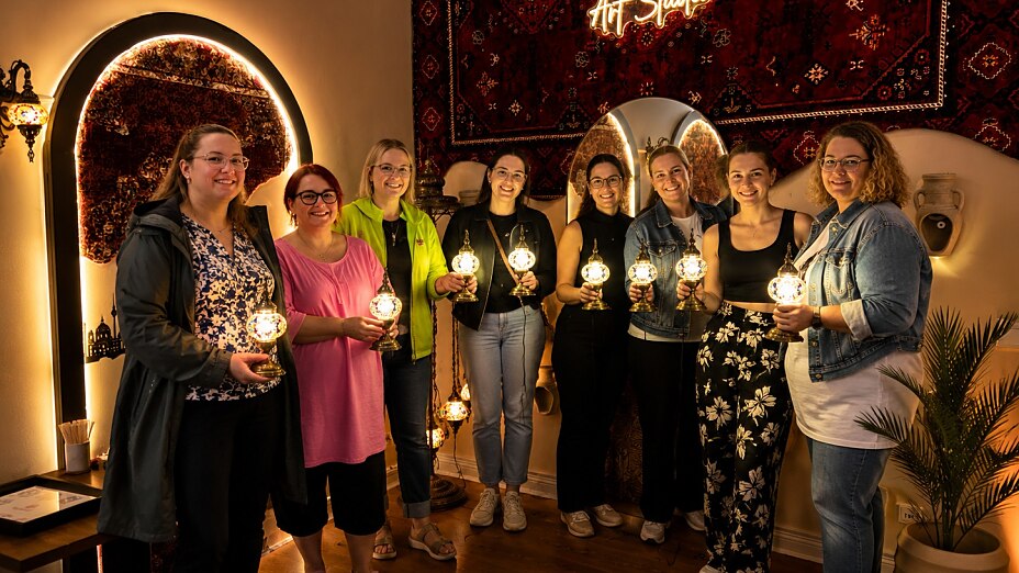 Frauen halten ihre selbst gestalteten leuchtenden Mosaiklampen während eines kreativen Workshops im Orient Art Studio Berlin.