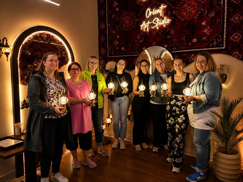 Frauen halten ihre selbst gestalteten leuchtenden Mosaiklampen während eines kreativen Workshops im Orient Art Studio Berlin.